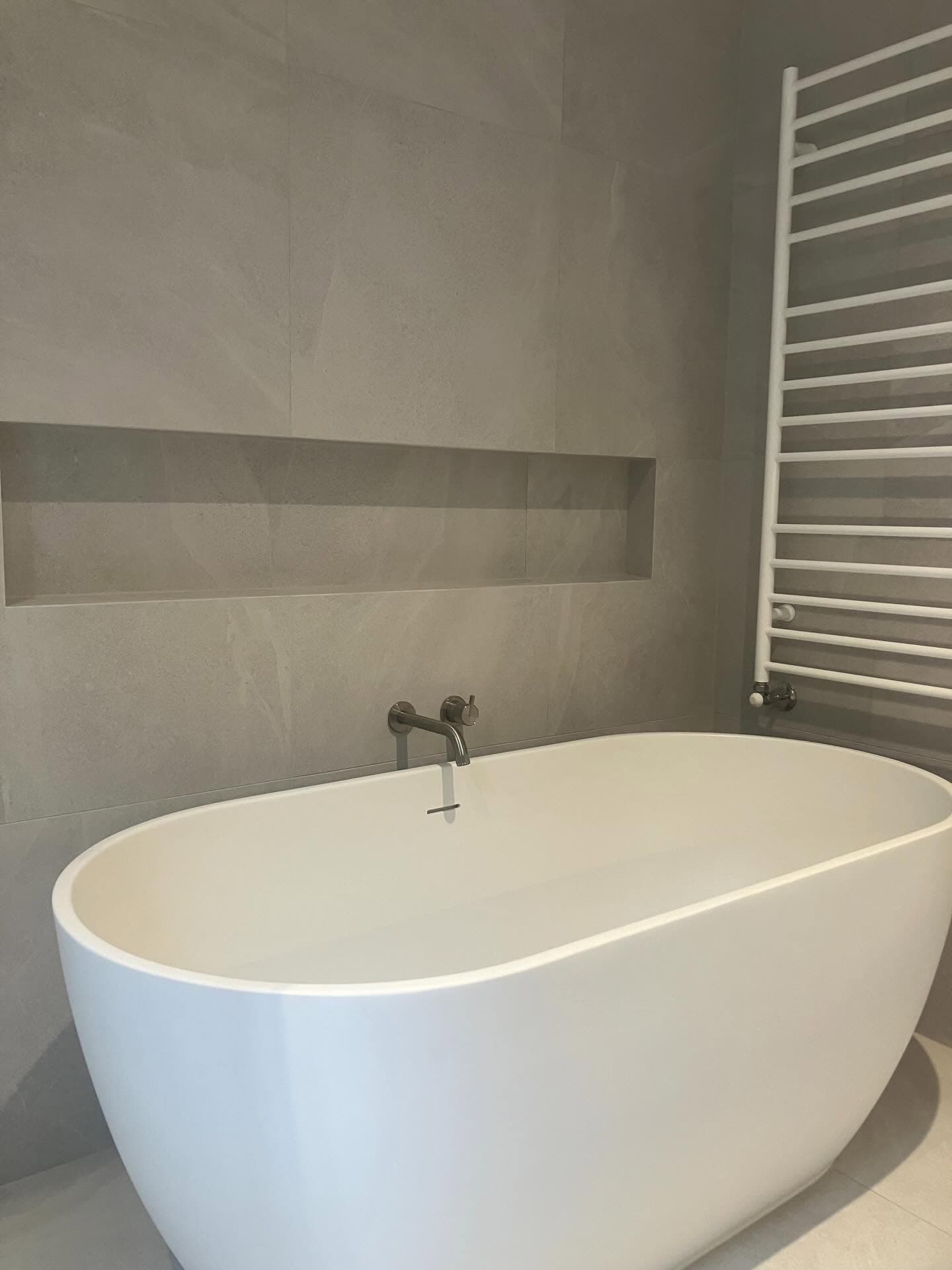 Freestanding white bath against a large-format stone wall, with a recessed shelf and a heated towel rail