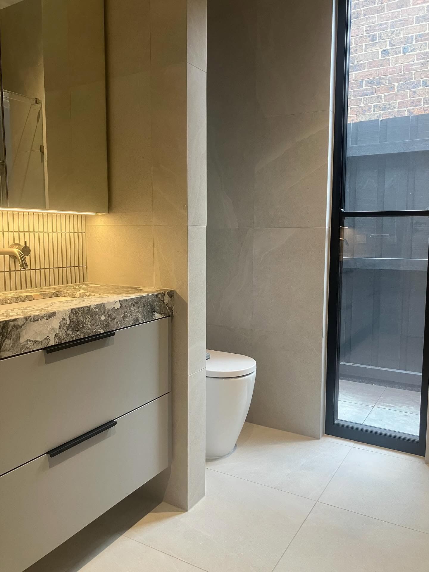 Warm beige wall tiles behind a stone vanity top and a wall-hung toilet, beside a black-framed shower