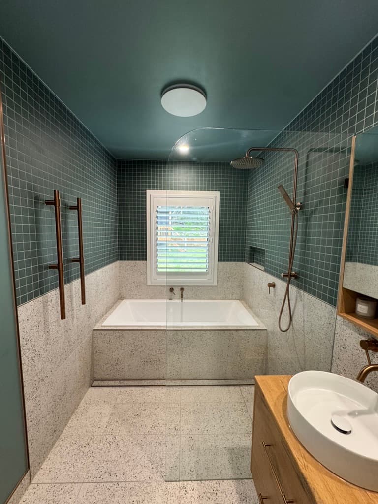 Bathroom in dark green square tiles over white terrazzo, with a built-in bath, a copper shower rail and a shuttered window