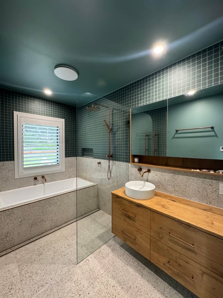 Oak vanity with a round white basin and copper tapware under a mirror cabinet, against dark green square tiles and white terrazzo