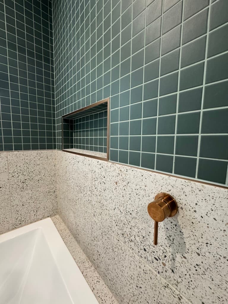 Dark green square tiles with a recessed niche above white terrazzo, and a copper mixer beside the bath