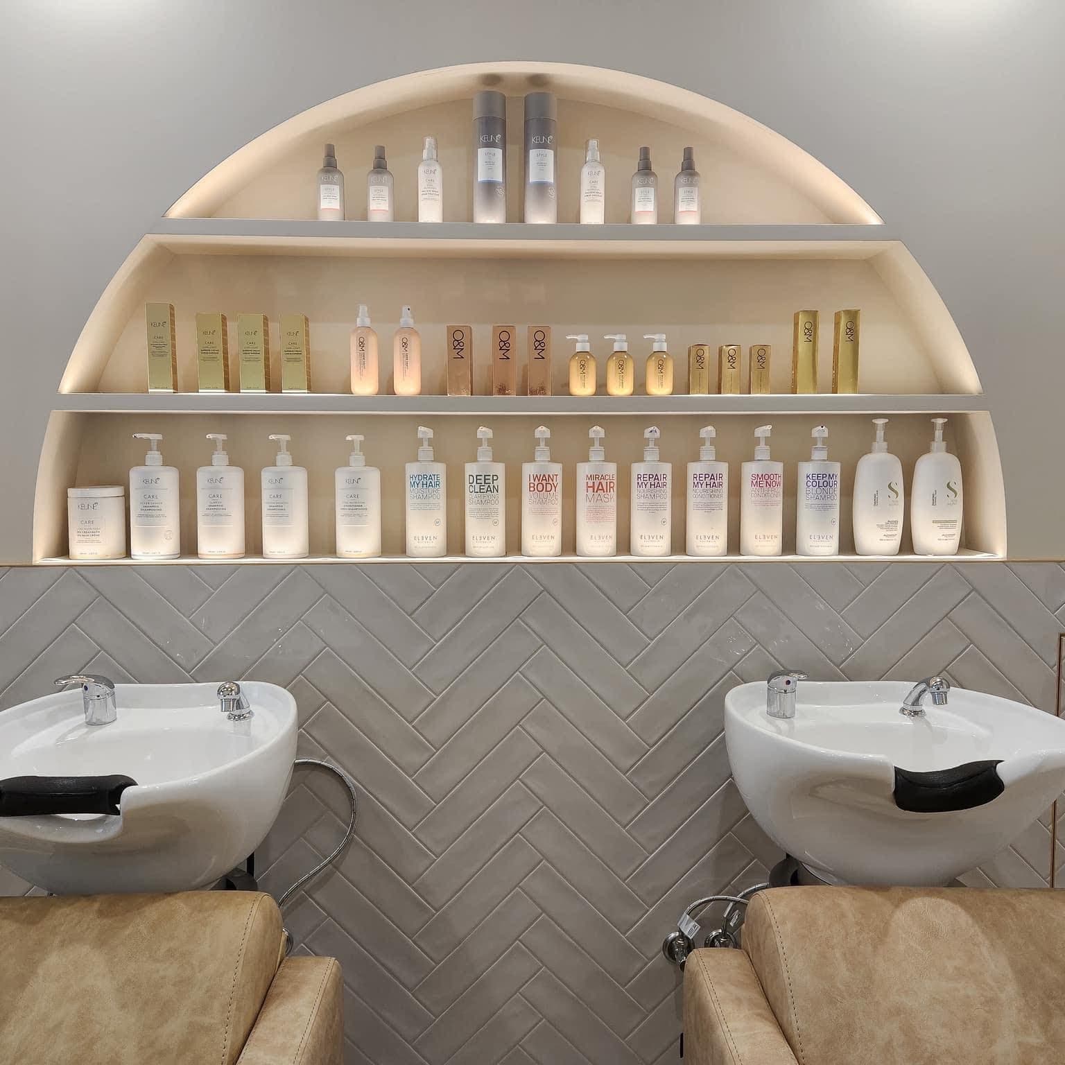 Salon basin wall tiled in pale herringbone, with an arched product niche above two backwash basins
