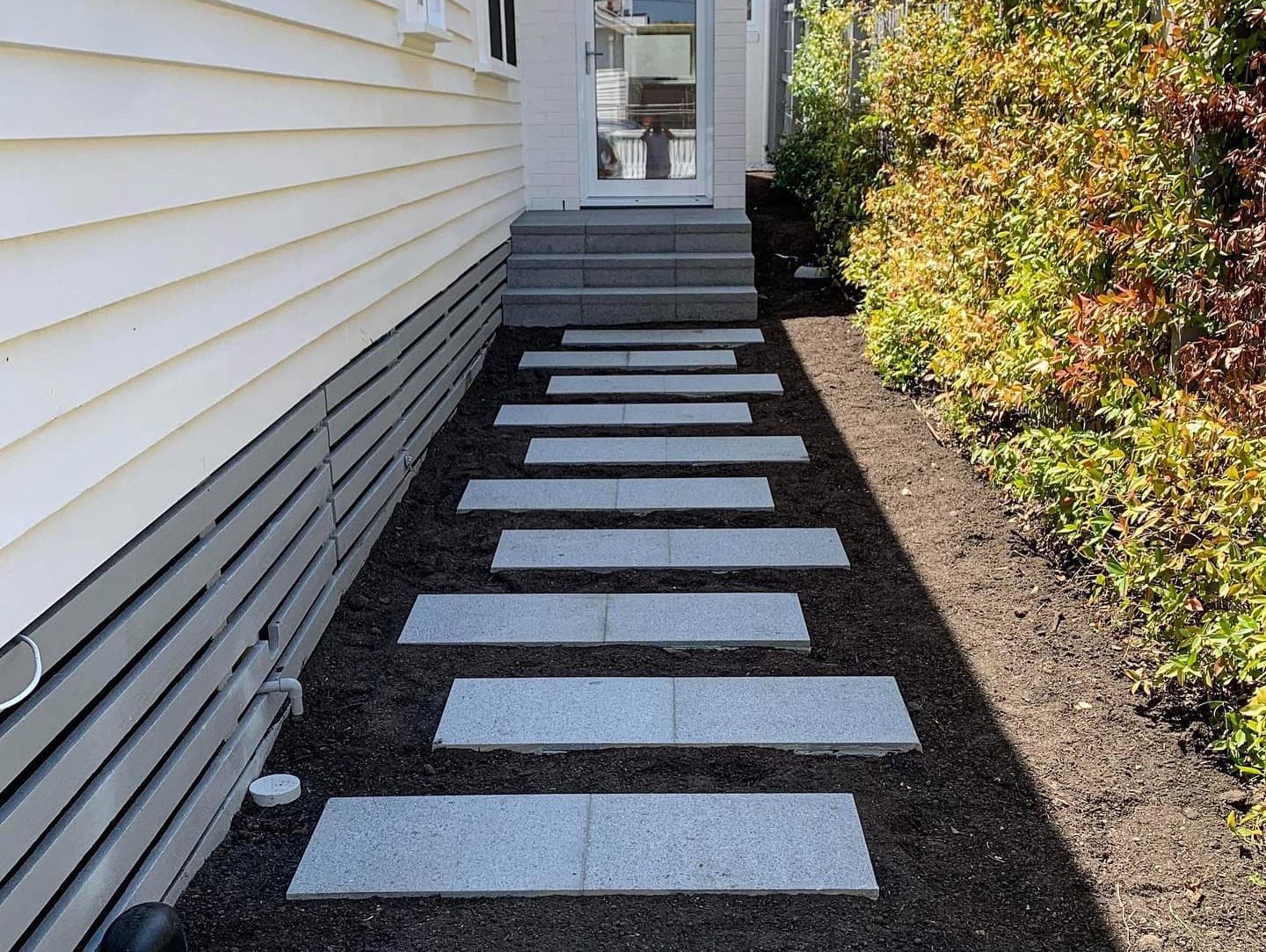 Rectangular stone stepping stones set into a garden bed along the side of a weatherboard house